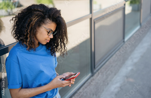 Beautiful African American woman wearing eyeglasses holding smartphone, text messaging on the street, copy space. Pensive female using mobile app shopping online