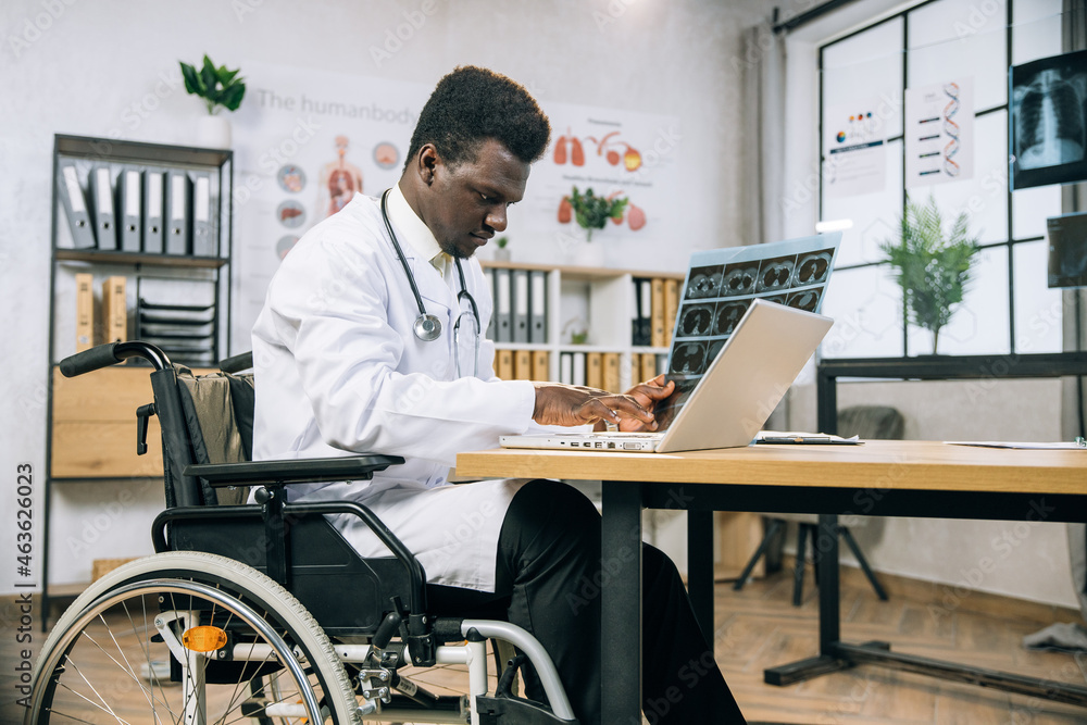 Focused african doctor with disability typing on laptop while analysing results of x ray scan. Medical specialist who using wheelchair working at bright modern cabinet. Focused african doctor with disability typing on laptop while analysing results of x ray scan. Medical specialist who using wheelchair working at bright modern cabinet.