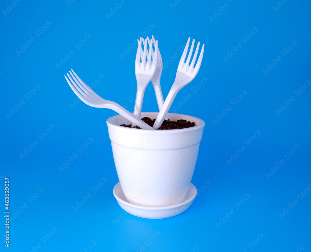 White pot with earth and plastic forks on a blue background. Plastic in the ground. Ecological problems White pot with earth and plastic forks on a blue background. Plastic in the ground. Ecological problems