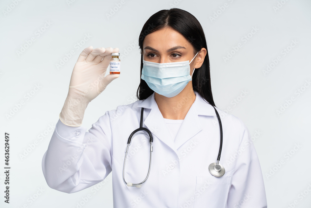 Medicine, profession and healthcare concept. Happy smiling female doctor wearing protective mask and white coat holding bottle with covid 19 vaccine over white background Medicine, profession and healthcare concept. Happy smiling female doctor wearing protective mask and white coat holding bottle with covid 19 vaccine over white background