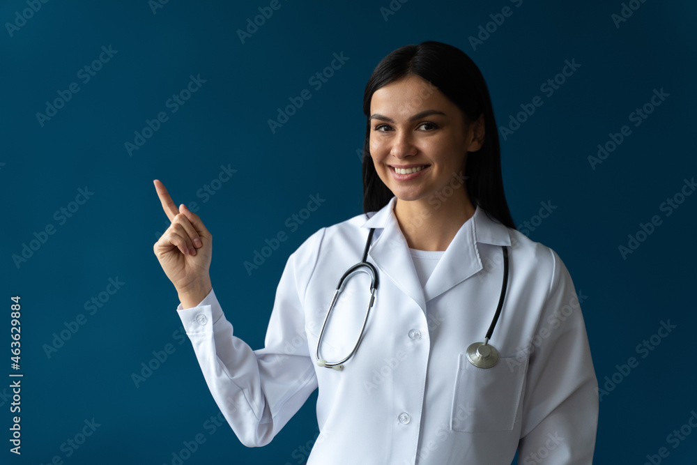 Waist up portrait view of the smiling happy doctor pointing with her finger at the free space on blue background. Doctor and medicine concept