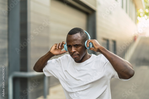 Chico negro atlético con cascos inalámbricos celestes escuchando música, sonriendo y bailando por la calle © MiguelAngelJunquera
