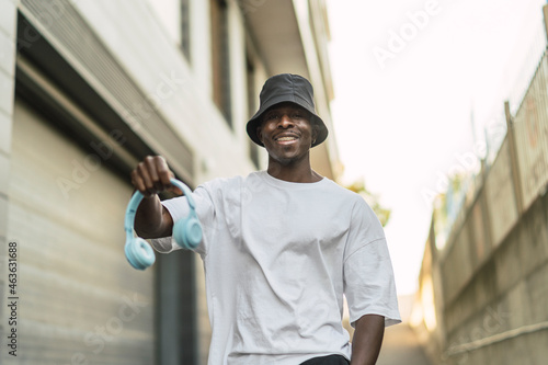 Chico negro atlético con cascos inalámbricos celestes escuchando música, sonriendo y bailando por la calle