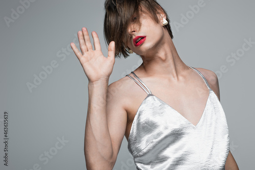 young transgender person with red lips and earring posing in slip dress isolated on gray