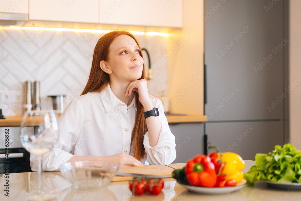 Medium shot portrait of dreaming redhead young woman sitting at kitchen table with cutting board, knife and fresh vegetables, looking up. Beautiful female cooking vegetarian meals at home. Medium shot portrait of dreaming redhead young woman sitting at kitchen table with cutting board, knife and fresh vegetables, looking up. Beautiful female cooking vegetarian meals at home.