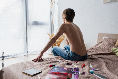 back view of shirtless transgender person sitting on bed near makeup products