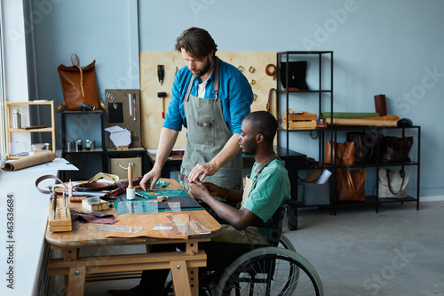 Side view portrait of young African-American man in wheelchair learning leathermaking craft in leatherworkers workshop, copy space photo