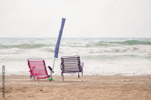 Fototapeta Naklejka Na Ścianę i Meble -  A couple of empty chairs on the beach with the umbrella closed on a cloudy day with the ocean in the background