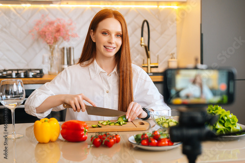 Medium shot portrait of smiling attractive redhead woman vlogger shooting video food blog about cooking on camera of mobile phone  sitting at kitchen with modern light interior  selective focus.