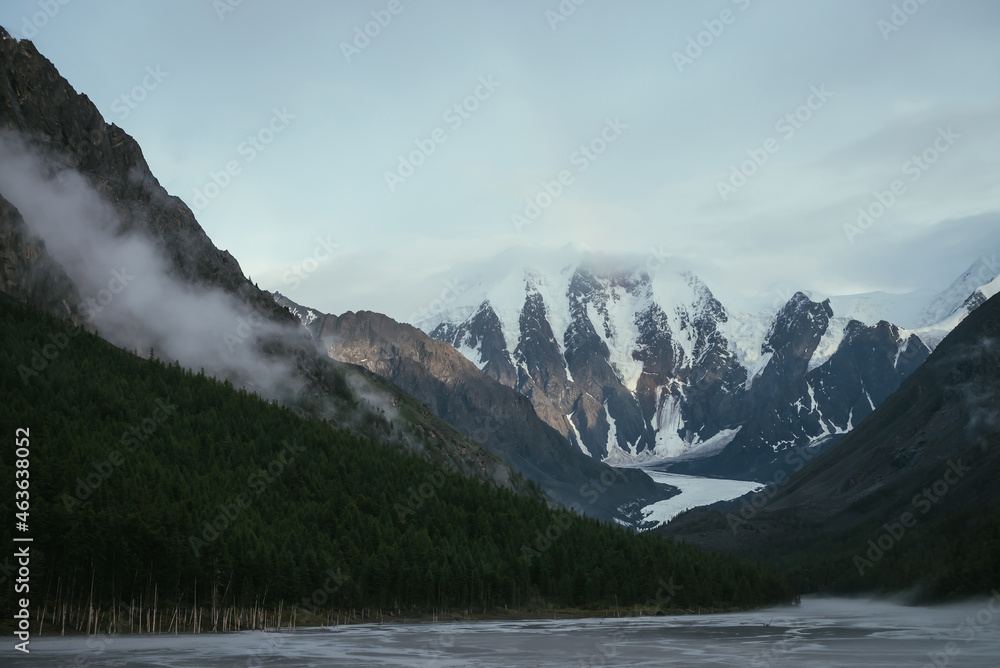 Scenic alpine landscape with mountain streams in fog and snowy mountains with rainbow under cloudy sky. Atmospheric scenery with shallow mountain lake, sunlight on rocks and low clouds in valley. Scenic alpine landscape with mountain streams in fog and snowy mountains with rainbow under cloudy sky. Atmospheric scenery with shallow mountain lake, sunlight on rocks and low clouds in valley.
