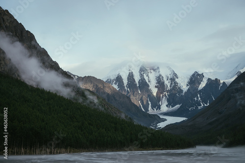 Scenic alpine landscape with mountain streams in fog and snowy mountains with rainbow under cloudy sky. Atmospheric scenery with shallow mountain lake, sunlight on rocks and low clouds in valley.