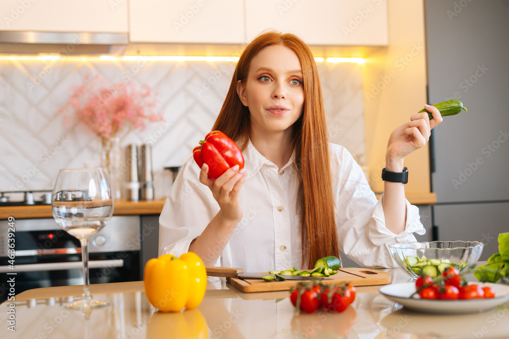 Medium shot portrait of cheerful redhead young woman holding in hand peppers sitting at wooden table with fresh vegetables, in light kitchen room with modern interior, looking away. Medium shot portrait of cheerful redhead young woman holding in hand peppers sitting at wooden table with fresh vegetables, in light kitchen room with modern interior, looking away.