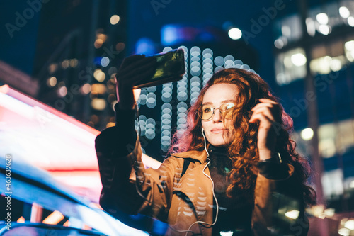 Curly haired young woman in glasses wearing light coat listens to music with headphones and makes selfie using phone camera against skyscraper in night city. photo