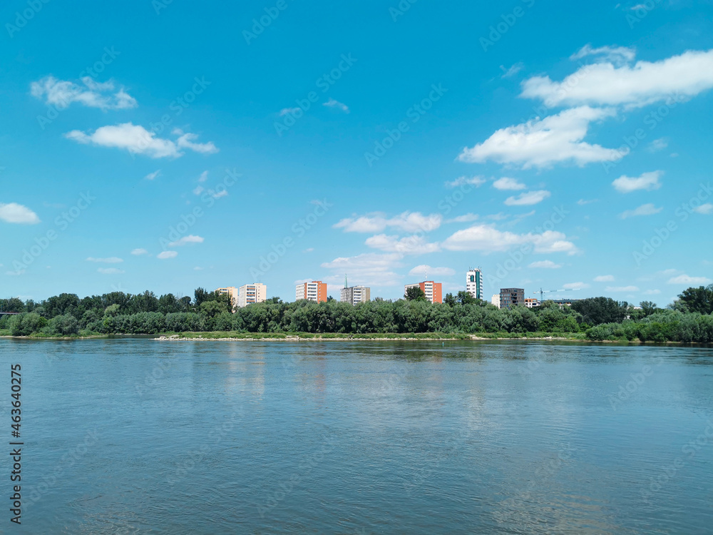 Beautiful summer or spring landscape of the river, blue sky with white clouds and protruding buildings among green trees