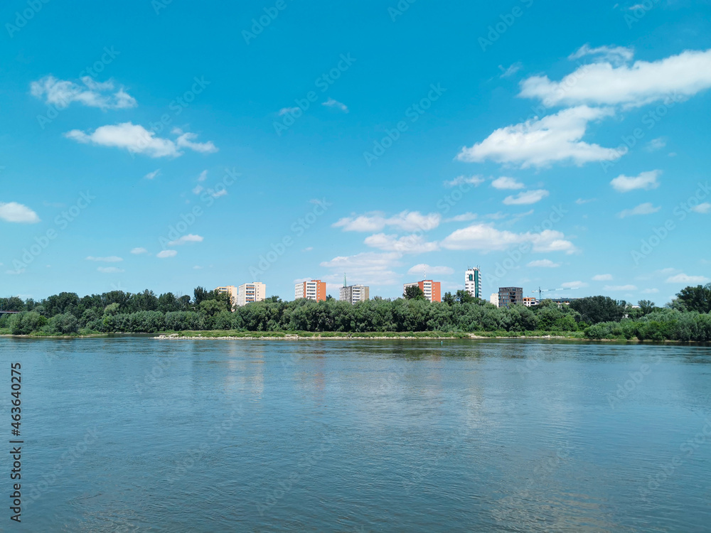 Beautiful summer or spring landscape of the river, blue sky with white clouds and protruding buildings among green trees Beautiful summer or spring landscape of the river, blue sky with white clouds and protruding buildings among green trees