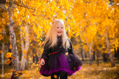 Little cute blonde girl dressed as a witch throws leaves in the park in autumn and smiles. Decorations for All Saints Day. Halloween costume.