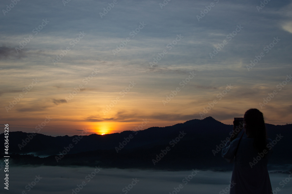 A dark silhouette of a tourist using a camera to capture the light of the morning sun.