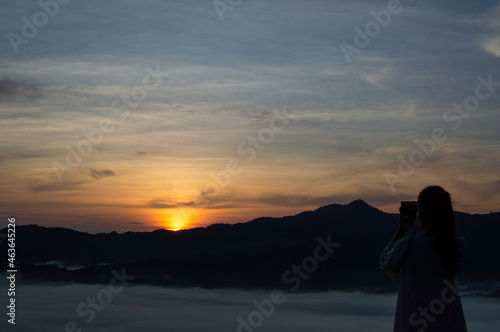 A dark silhouette of a tourist using a camera to capture the light of the morning sun. © wittaya