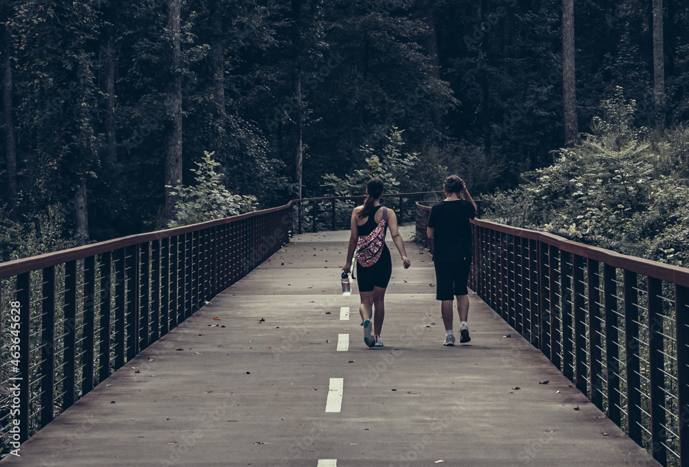 two people walking across pedestrian bridge on walking trail surrounded by forest in background