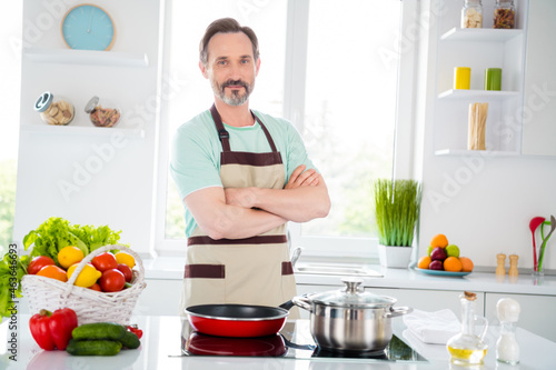Photo of self-assured man prepare pan folded arms posing wear apron blue t-shirt home kitchen indoors