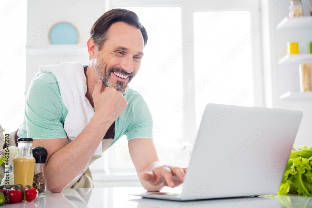 Photo of blogger man typing netbook read news recipe wear kitchenware apron in kitchen apartment indoors Photo of blogger man typing netbook read news recipe wear kitchenware apron in kitchen apartment indoors