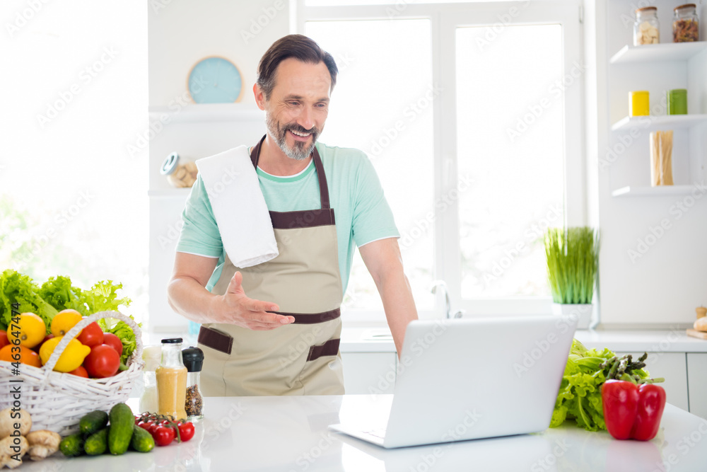 Photo of confident nice man look screen laptop video call lesson wear apron blue t-shirt home kitchen indoors Photo of confident nice man look screen laptop video call lesson wear apron blue t-shirt home kitchen indoors