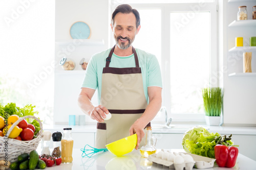 Photo of cheerful positive mature guy wear blue t-shirt apron cooking omelet smiling indoors house room