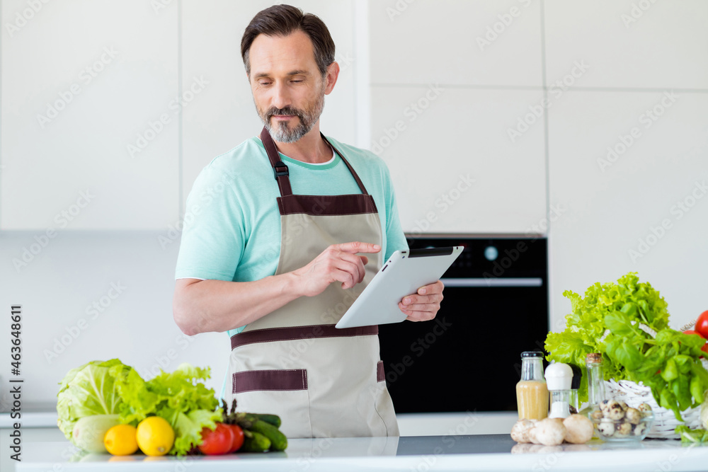 Photo of serious chef man hold e-book read recipe cook salad wear kitchenware apron in kitchen apartment indoors Photo of serious chef man hold e-book read recipe cook salad wear kitchenware apron in kitchen apartment indoors