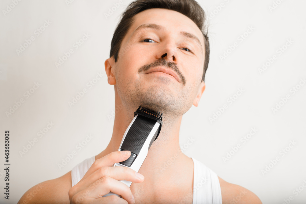 Portrait of handsome unshaven man shaving modern electric razor and looking at camera indoors. Selective focus on beard and mustache grooming device, trimmer. Men's beauty, daily skin care Portrait of handsome unshaven man shaving modern electric razor and looking at camera indoors. Selective focus on beard and mustache grooming device, trimmer. Men's beauty, daily skin care