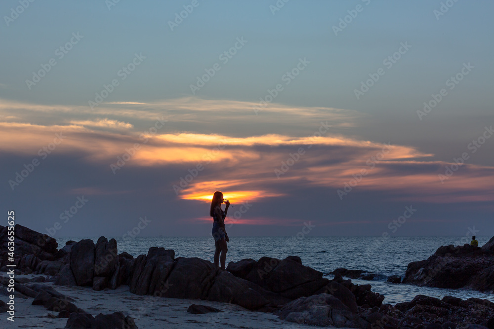 Woman standing on the rocks by the sea holding a wine glass. and playing mobile phone during sunset at Patong Beach, Phuket, Thailand. Woman standing on the rocks by the sea holding a wine glass. and playing mobile phone during sunset at Patong Beach, Phuket, Thailand.