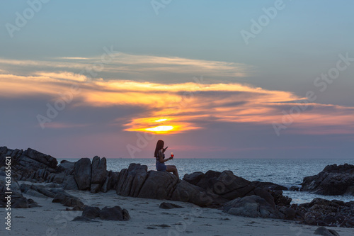 Photography A lady sitting on the rocks by the sea holding a wine glass and playing mobile phone during sunset at Patong Beach, Phuket, Thailand