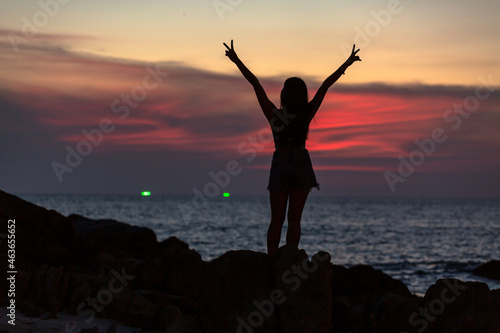 Woman standing waving on the rocks by the sea in the red sky at twilight at Patong Beach, Phuket, Thailand..