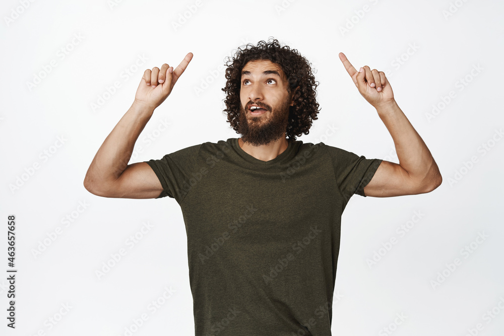 Image of young curly guy pointing fingers up, looking amazed by sale offer, standing in t-shirt over white background Image of young curly guy pointing fingers up, looking amazed by sale offer, standing in t-shirt over white background