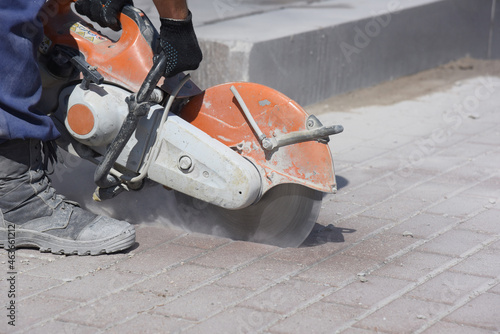 Close-up of a worker in overalls with a circular saw cutting paving concrete tiles laid out in even rows.