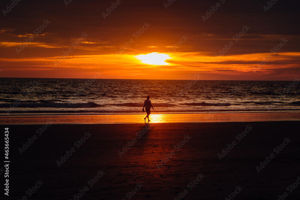 man walking calmly on the beach reflected in the shore with intense sunset of orange and purple colors with soft clouds