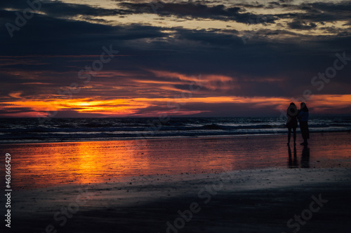 couple of women standing on the shore of the beach watching the intense sunset of orange and purple colors with soft clouds