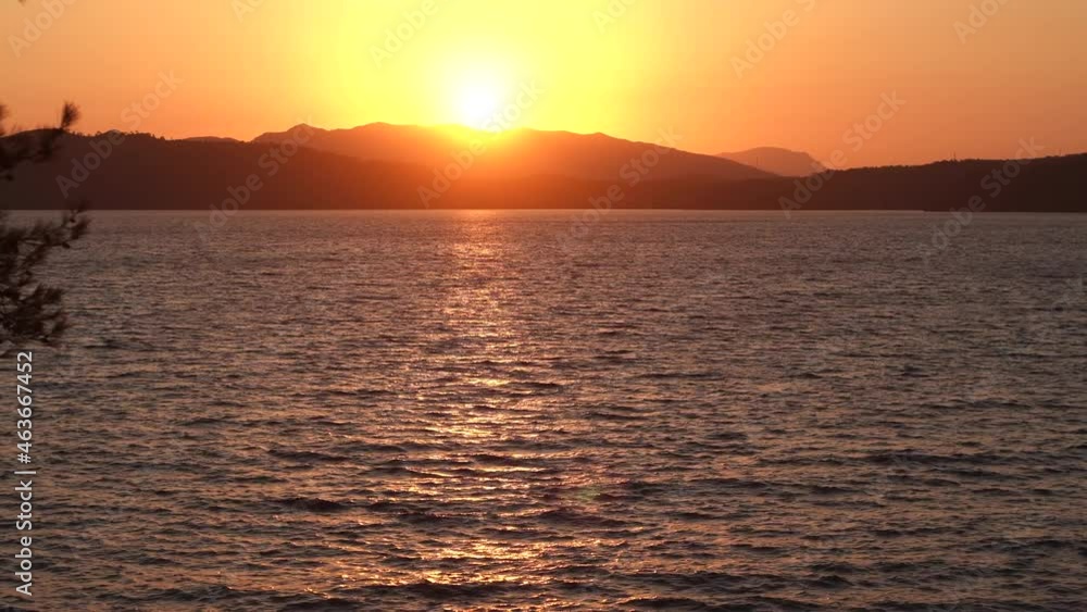 Beautiful sunset golden peaceful marine landscape. 4k video footage of orange sky, twilight sun over tops of mountains, sea water, dark silhouettes of branches of tree growing on shore