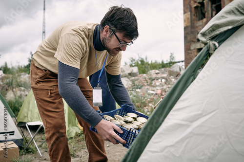 Male volunteer with box of canned food bending by tent where migrants live