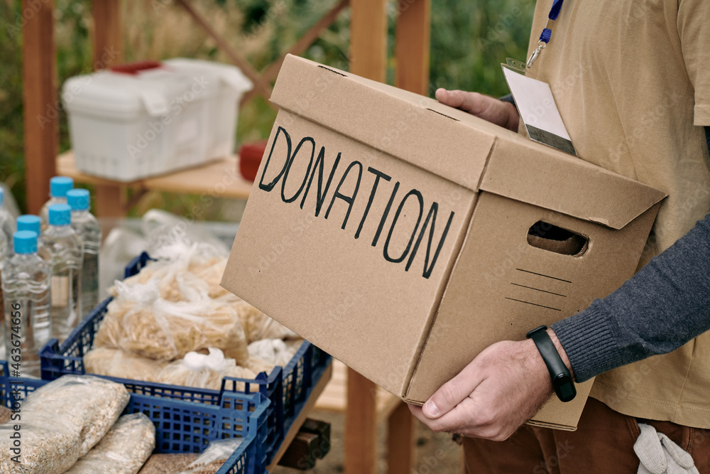 Male volunteer holding packed donation box while standing by table with free food - obrazy, fototapety, plakaty 
