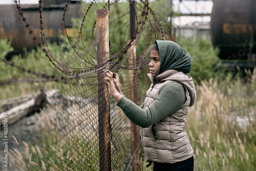 Homeless and lonely girl in casualwear standing by barb wire fence on the territory of refugee camp photo