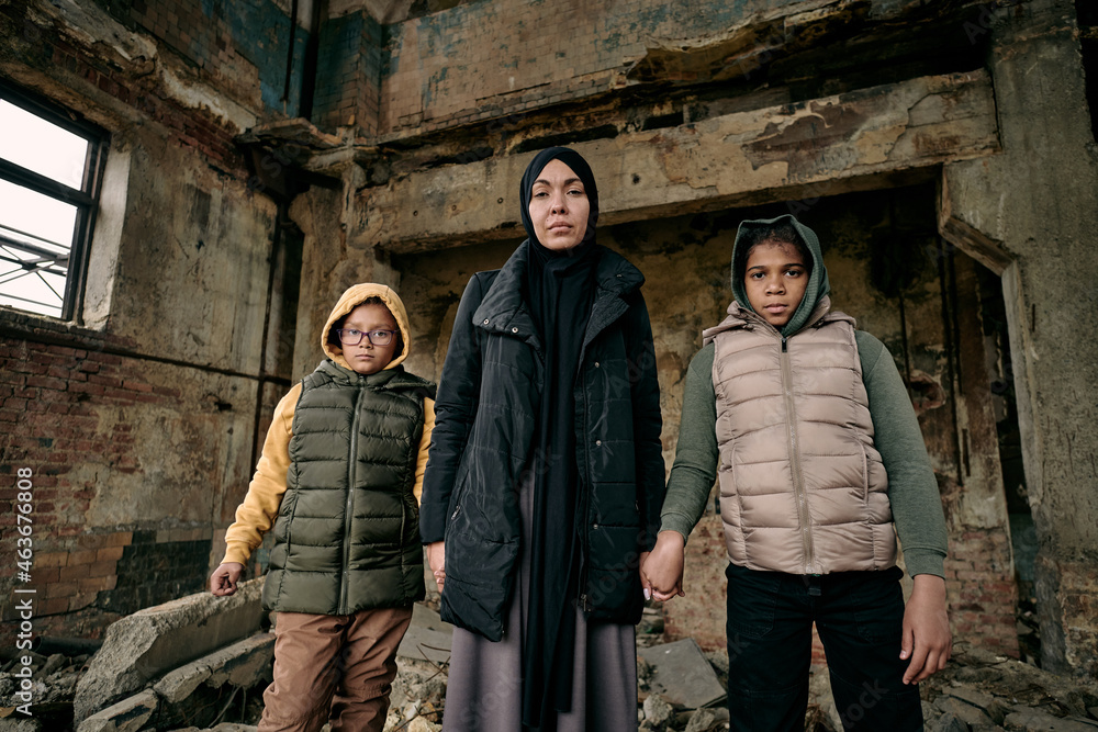 Young Muslim woman holding her two daughters by hands while standing in half ruined house Young Muslim woman holding her two daughters by hands while standing in half ruined house