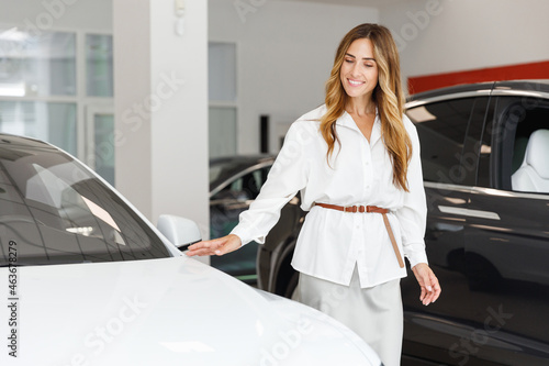 Happy smiling fun woman customer female buyer client in white shirt touch hood choose auto want to buy new automobile in car showroom vehicle salon dealership store motor show indoor. Sales concept. © ViDi Studio
