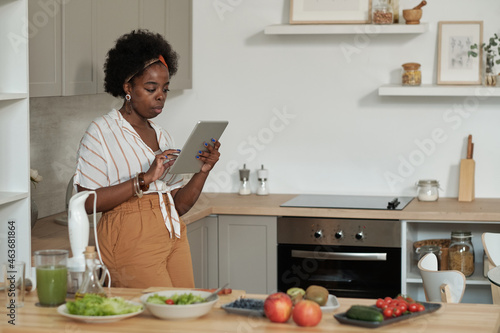 Young African housewife with tablet looking through online recipes while standing in the kitchen