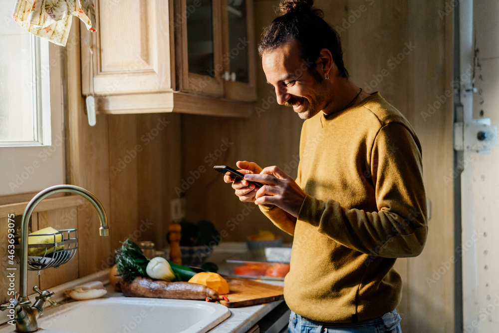 Young man looking at phone in kitchen while cooking Young man looking at phone in kitchen while cooking