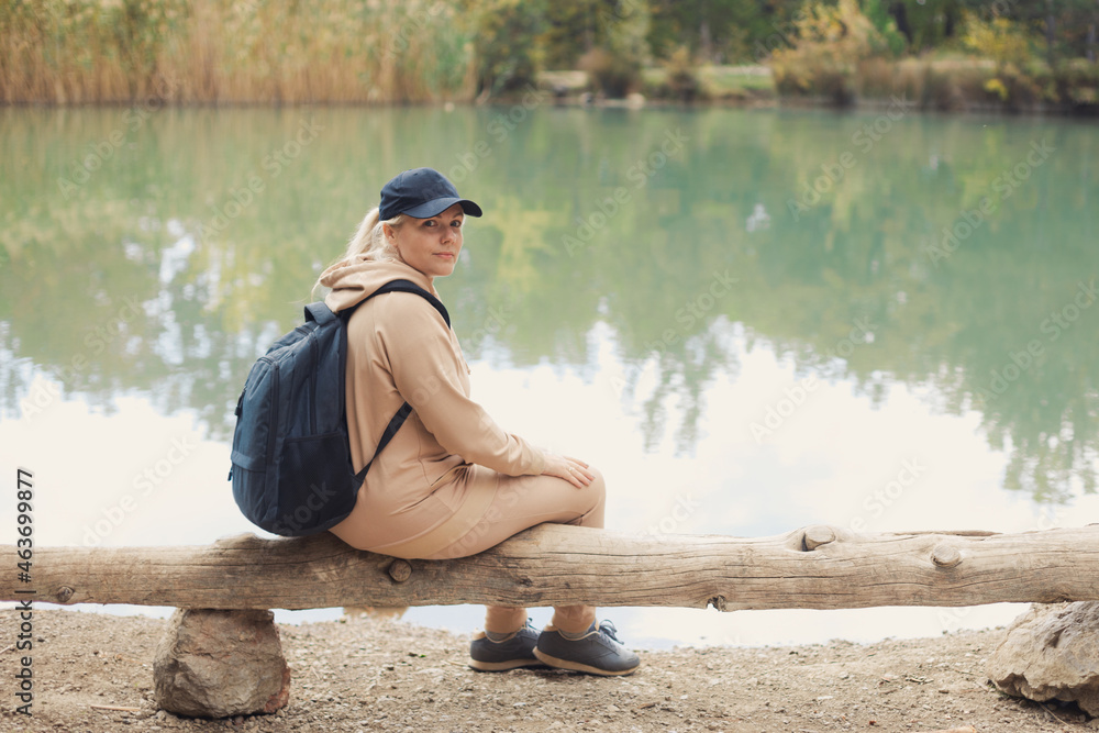 Full body portrait of caucasian plus size woman 30-35 years old with backpack in beige sportswear looking at camera, sitting on a log at a mountain lake, walking in the fall woods on vacations.