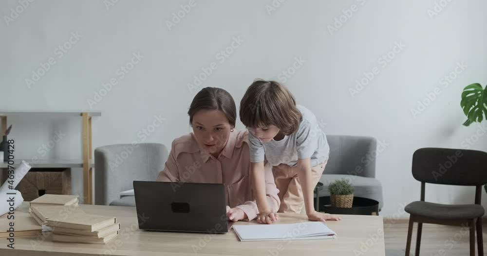 Freelance mom is focused on typing and working remotely at home on laptop. Restless son climbs up on the table and tries to touch keyboard, disturbing and annoying his mother. Freelance mom is focused on typing and working remotely at home on laptop. Restless son climbs up on the table and tries to touch keyboard, disturbing and annoying his mother.
