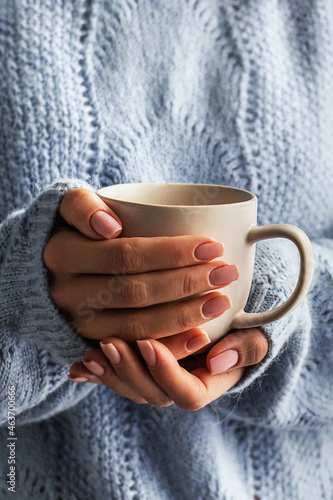 Female hands with a mug of drink close-up. Beautiful girl in a blue sweater holds a cup of tea or coffee in the morning sunlight. Autumn winter concept.