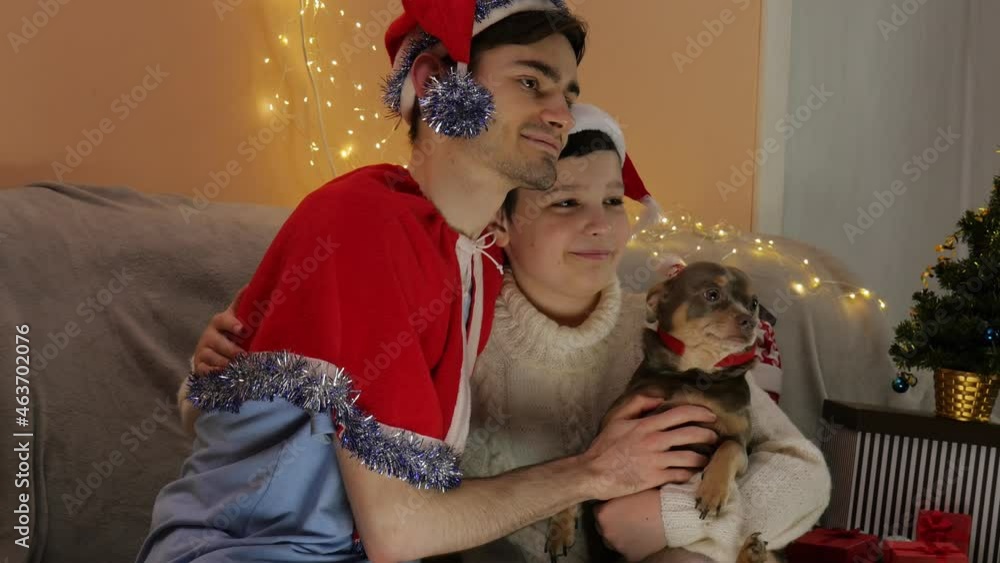 Two happy guys are celebrating Christmas. Santa Claus hat. Shining lights and a dog. Take a photo Two happy guys are celebrating Christmas. Santa Claus hat. Shining lights and a dog. Take a photo
