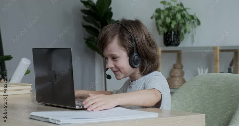 Enthusiastic cute boy video chatting merrily using headset and laptop with friends and parents and telling funny stories. Distance learning for kids in a playful way. Enthusiastic cute boy video chatting merrily using headset and laptop with friends and parents and telling funny stories. Distance learning for kids in a playful way.