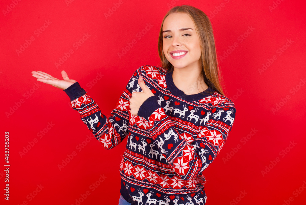 Young caucasian girl wearing christmas sweaters on red background Showing palm hand and doing ok gesture with thumbs up, smiling happy and cheerful. Young caucasian girl wearing christmas sweaters on red background Showing palm hand and doing ok gesture with thumbs up, smiling happy and cheerful.